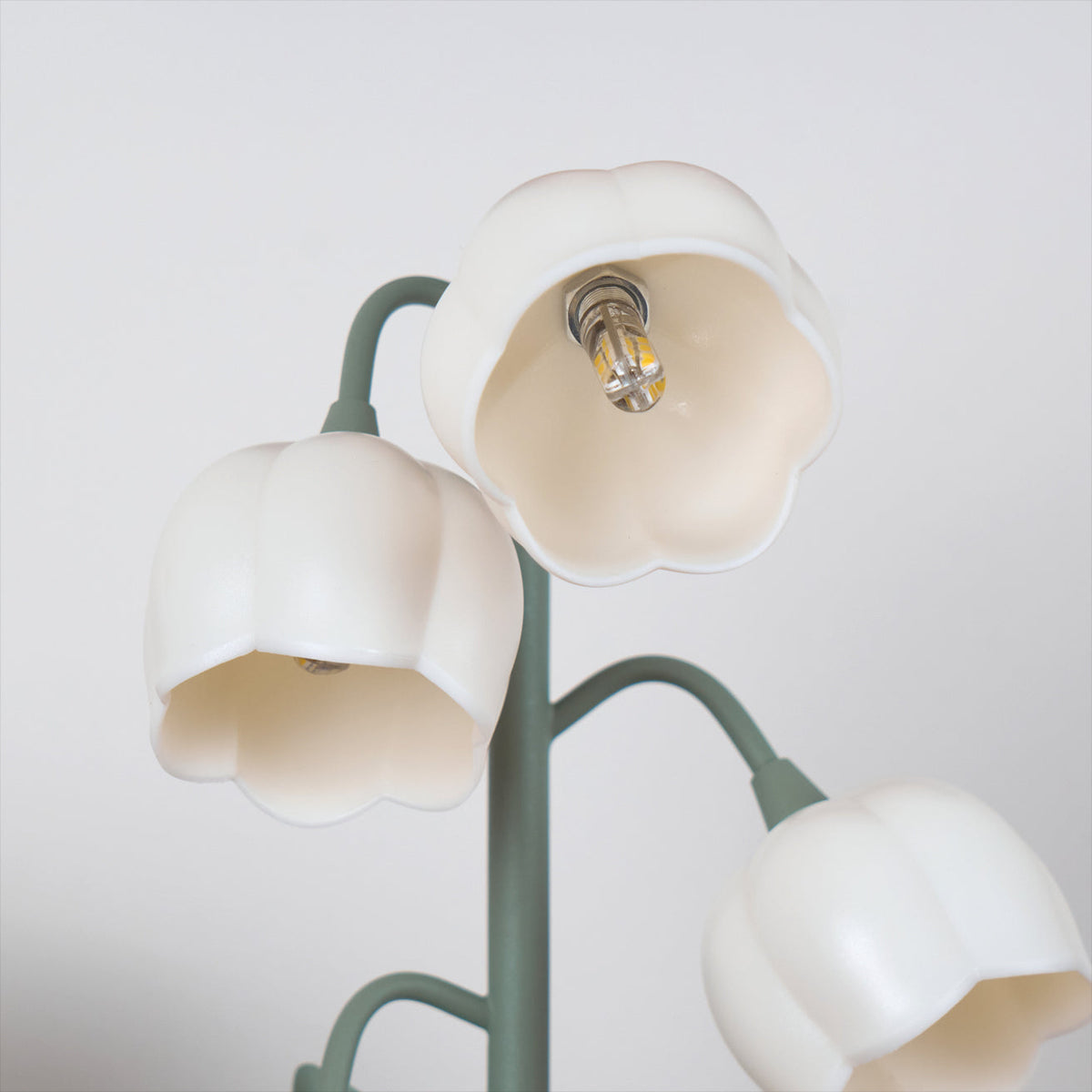 Close view of three white bell shades with the bulb visible inside one, on the slim green stems of this Clérachic table lamp.