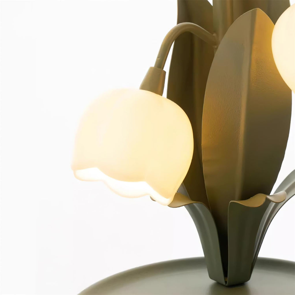 Close view of a lit bell shade on this Clérachic table lamp, with the green leaf blades and the curved stem beside it.
