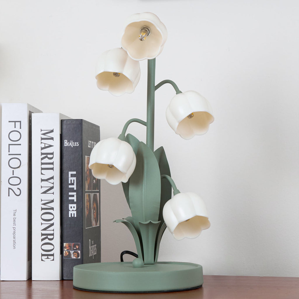 Clérachic table lamp, unlit, standing next to a row of upright books on a wooden shelf in front of a white wall.