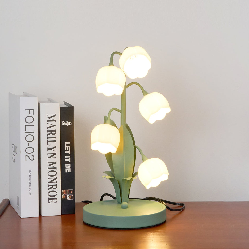 A Clérachic table lamp lit on a wooden shelf beside books titled with film and music names, in front of a white wall.