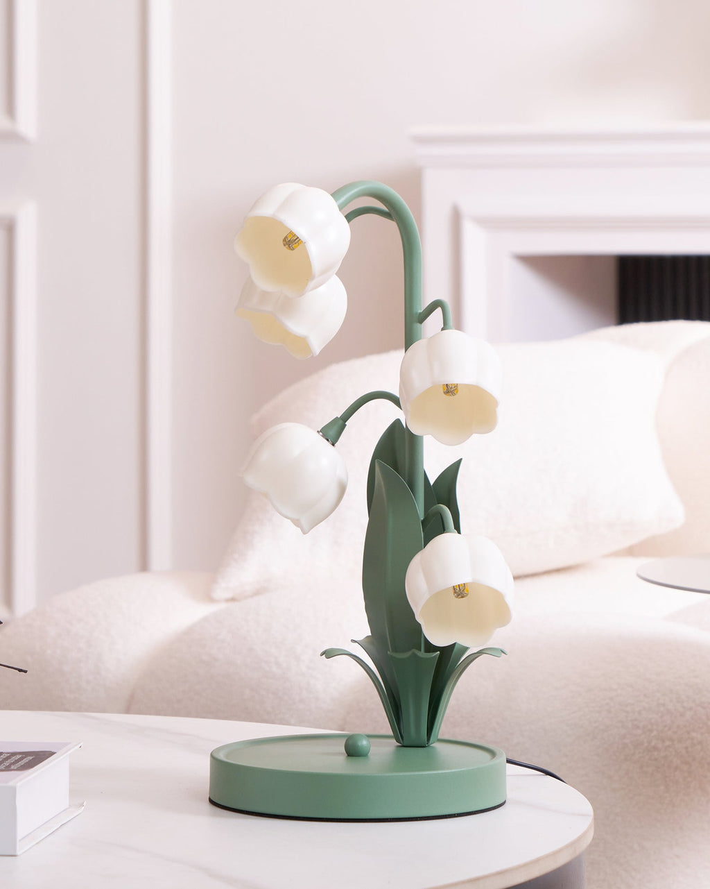A Clérachic table lamp, unlit, on a white marble table in front of a cream sofa and a white mantel.