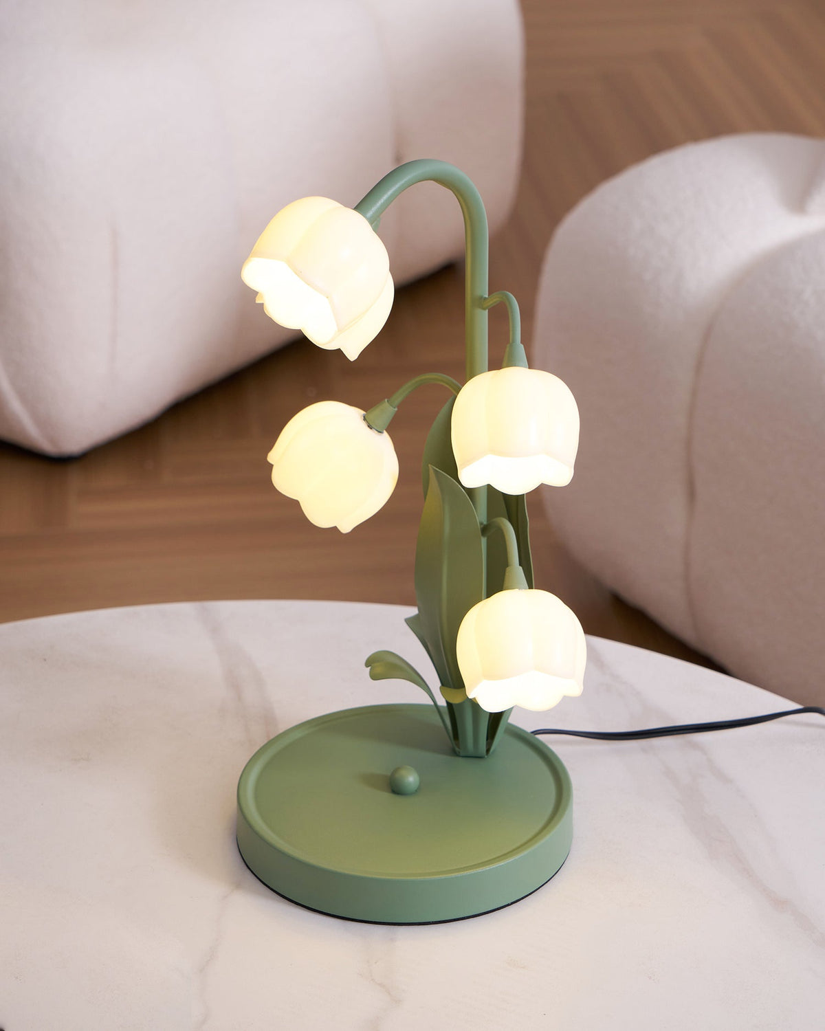 A lit Clérachic table lamp on a marble tray table, with a woven cream armchair and a wooden floor around it.