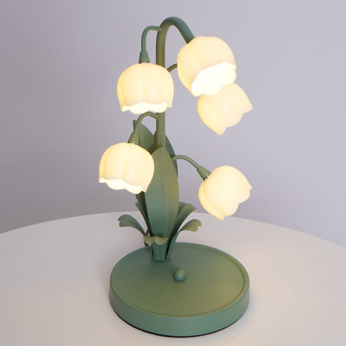 A lit Clérachic table lamp on a round white table, seen from above, with its green base and leaf blades.