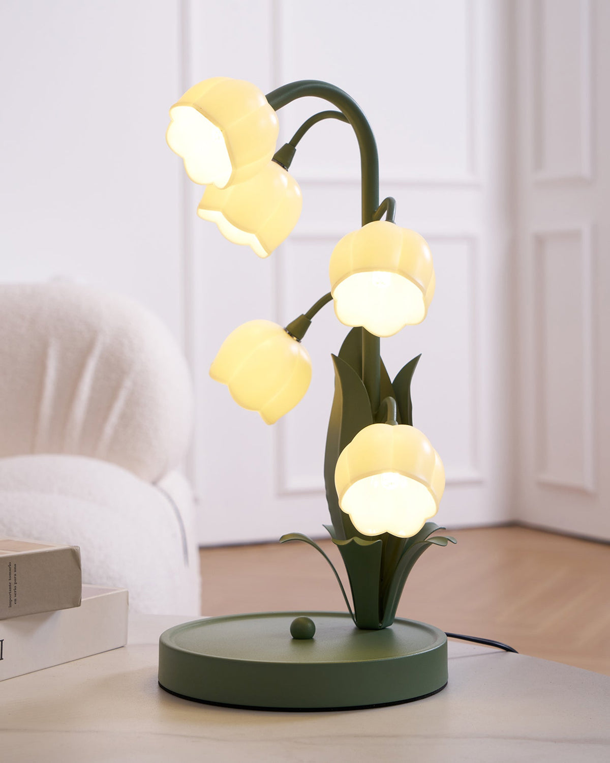 This Clérachic table lamp lit on a table beside a book and an armchair, with white panelled doors behind it.