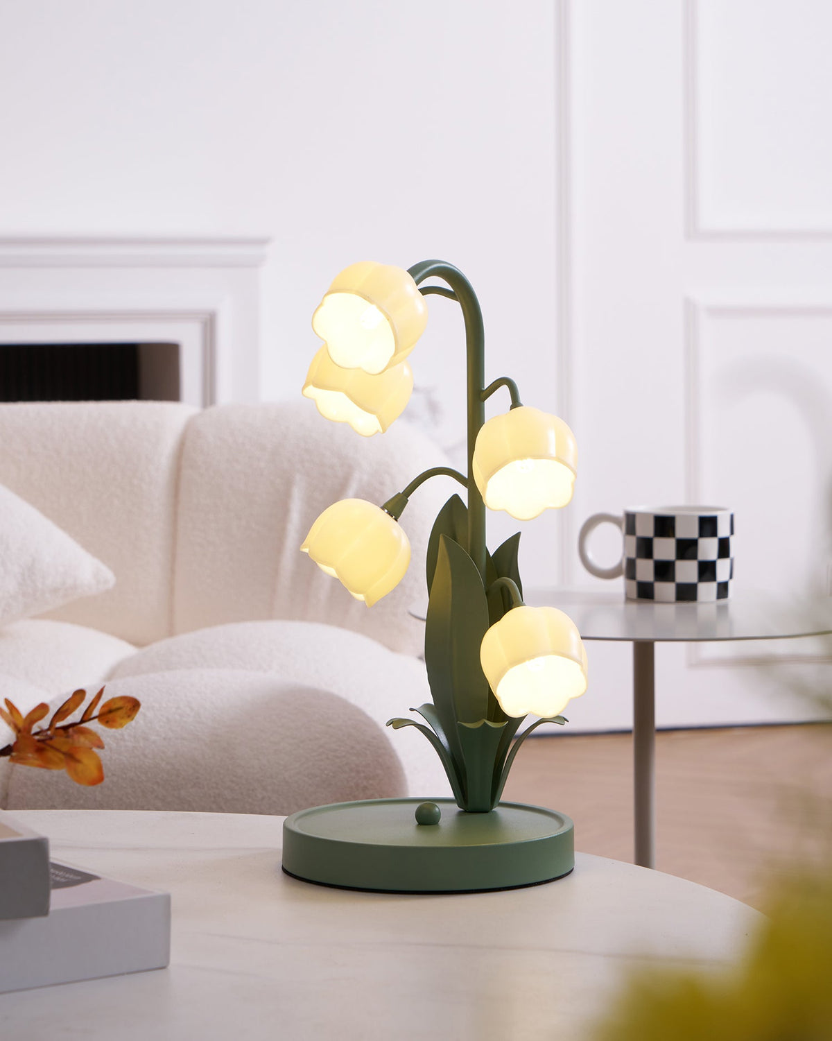 A lit Clérachic table lamp on a white table near a cream sofa, with a black and white mug on a glass side table behind it.