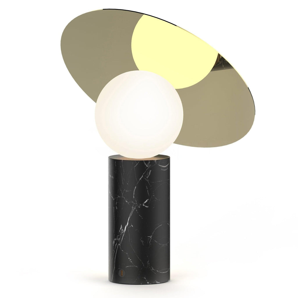 Clérachic disc table lamp lit on a plain white background, its black marble base topped by an olive-gold tilted disc.