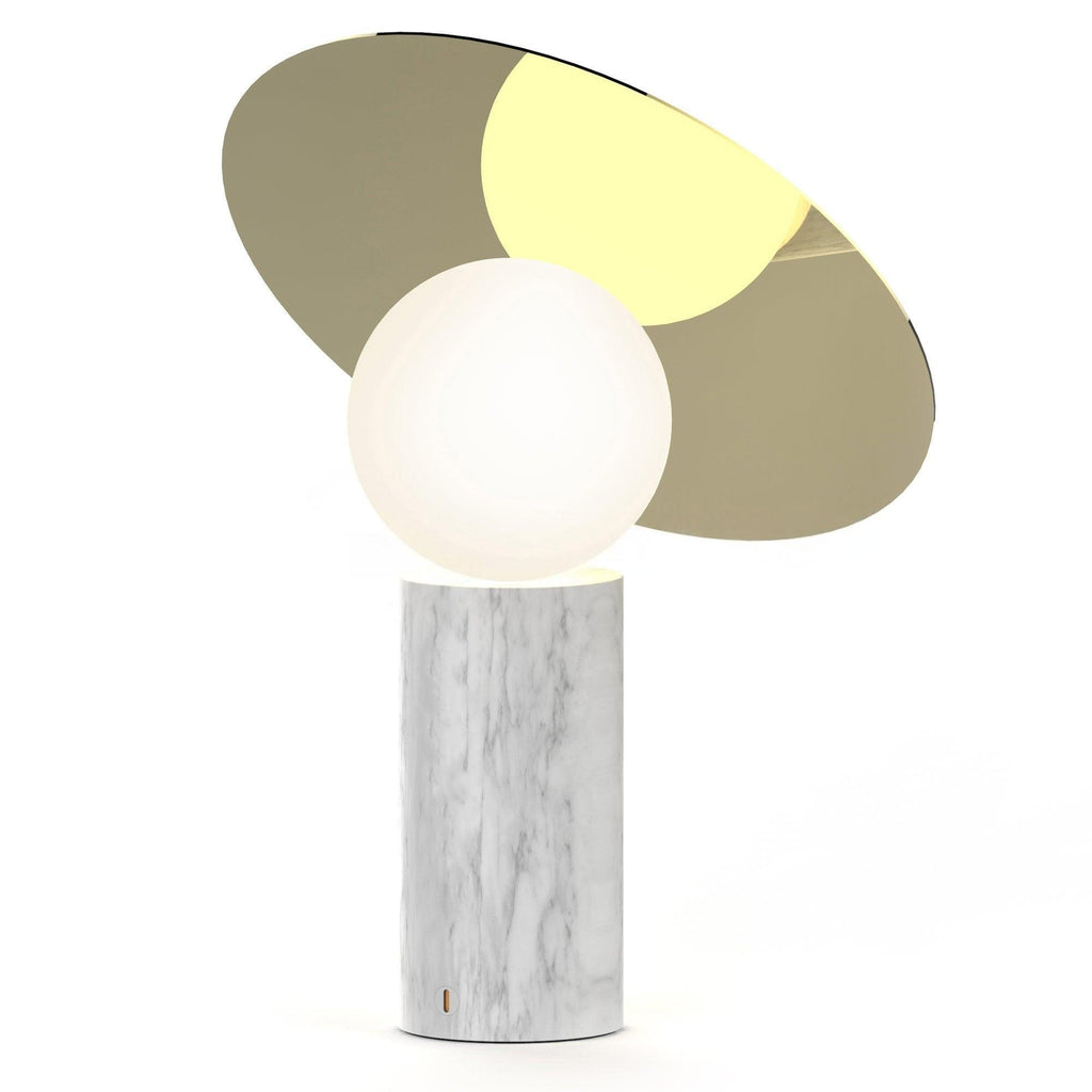 Clérachic disc lamp lit on a plain white background, its cylindrical white marble base topped by a warm gold-toned tilted disc.