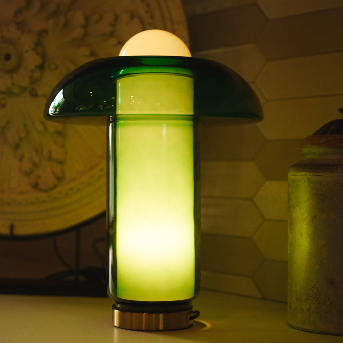 Clérachic green glass table lamp lit warm on a brushed brass base, with a white dome and the cylinder glowing lime green.