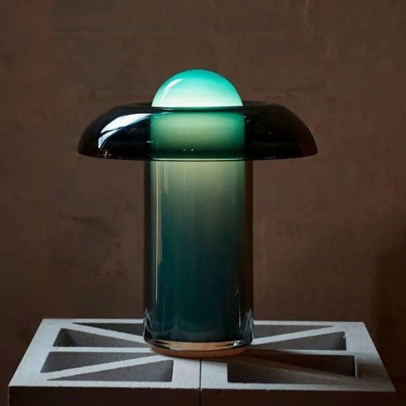 Clérachic green glass table lamp lit on a concrete breeze block, its dome glowing green against a dark terracotta wall.