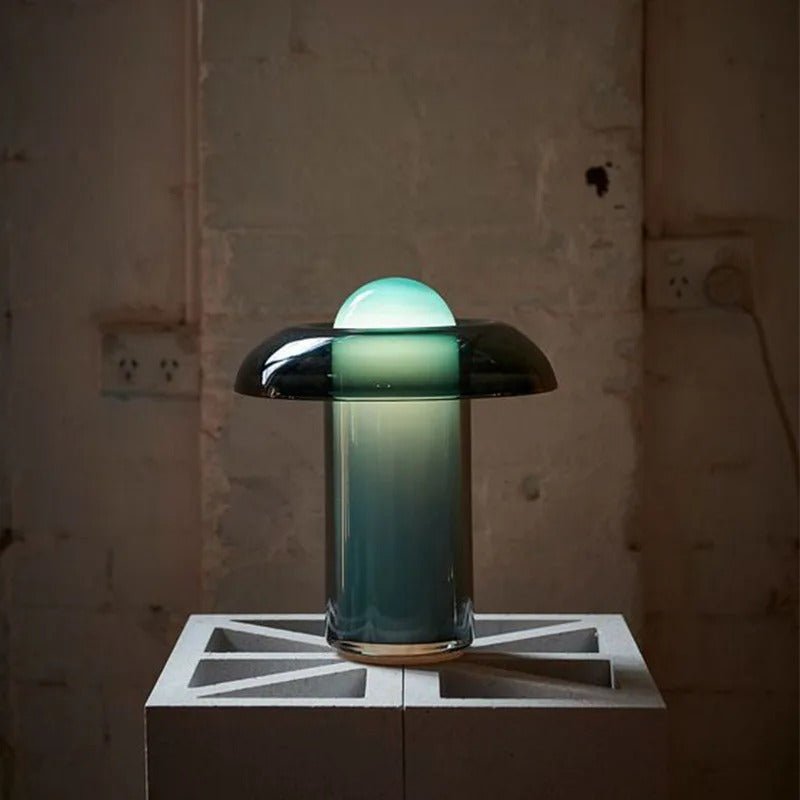 Clérachic green glass table lamp glowing on a concrete block against a worn, pale wall with two wall sockets.