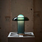 Clérachic green glass table lamp glowing on a concrete block against a worn, pale wall with two wall sockets.