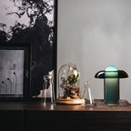 Clérachic green glass table lamp on a dark wood sideboard beside a glass cloche, flasks and a framed print.