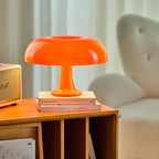 A Clérachic table lamp with a glossy orange dome shade, set on a stack of books on a warm wooden shelf beside a cream armchair.