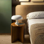 Small white dome lamp from Clérachic on a round wooden bedside table, next to a bed with grey bedding.