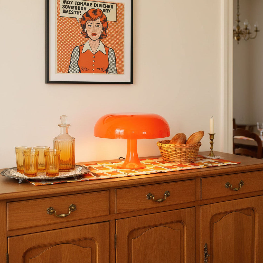A Clérachic orange table lamp glowing on a wooden sideboard, with amber glasses and a patterned runner beside it.