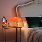 Orange dome lamp from Clérachic lit on a glass bedside table beside a bed with a green cushion and knitted throw.
