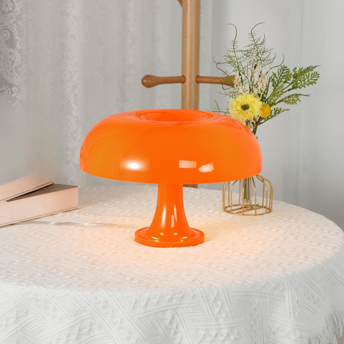 Glossy orange Clérachic table lamp on a table with a white lace cloth, with a small vase of yellow flowers behind.