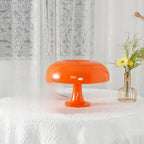 A Clérachic orange table lamp on a lace-covered table, its shade shining and a slim vase of yellow flowers to one side.