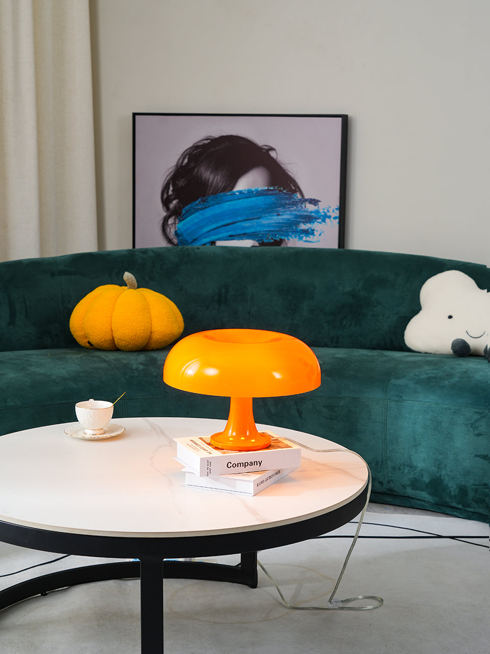 Warm-lit orange Clérachic table lamp on a round white coffee table in front of a dark green curved sofa.