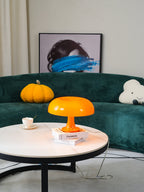 Warm-lit orange Clérachic table lamp on a round white coffee table in front of a dark green curved sofa.
