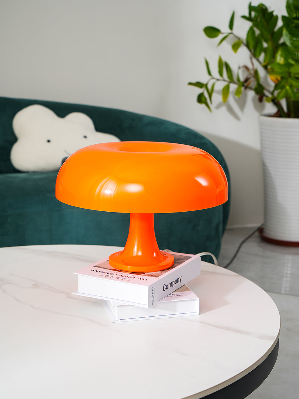 This Clérachic orange mushroom lamp stands on a pair of books on a white table, a green sofa and plant behind.