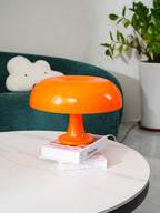 This Clérachic orange mushroom lamp stands on a pair of books on a white table, a green sofa and plant behind.