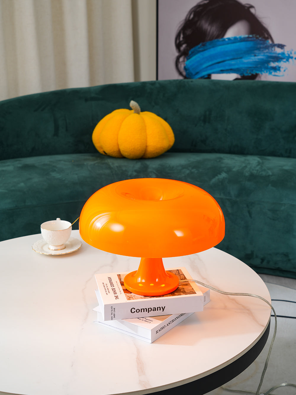 A Clérachic orange dome lamp on a stack of books on a round marble-top table, with a teacup and a green sofa behind.