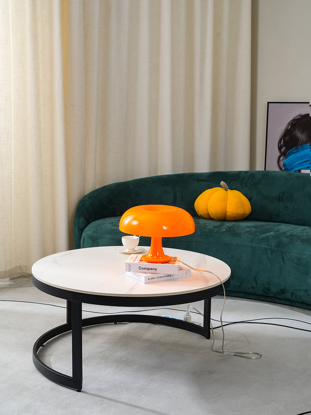 Wider view of this Clérachic orange lamp on a round table with a black base, in a room with cream curtains and a green sofa.
