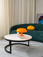 Wider view of this Clérachic orange lamp on a round table with a black base, in a room with cream curtains and a green sofa.