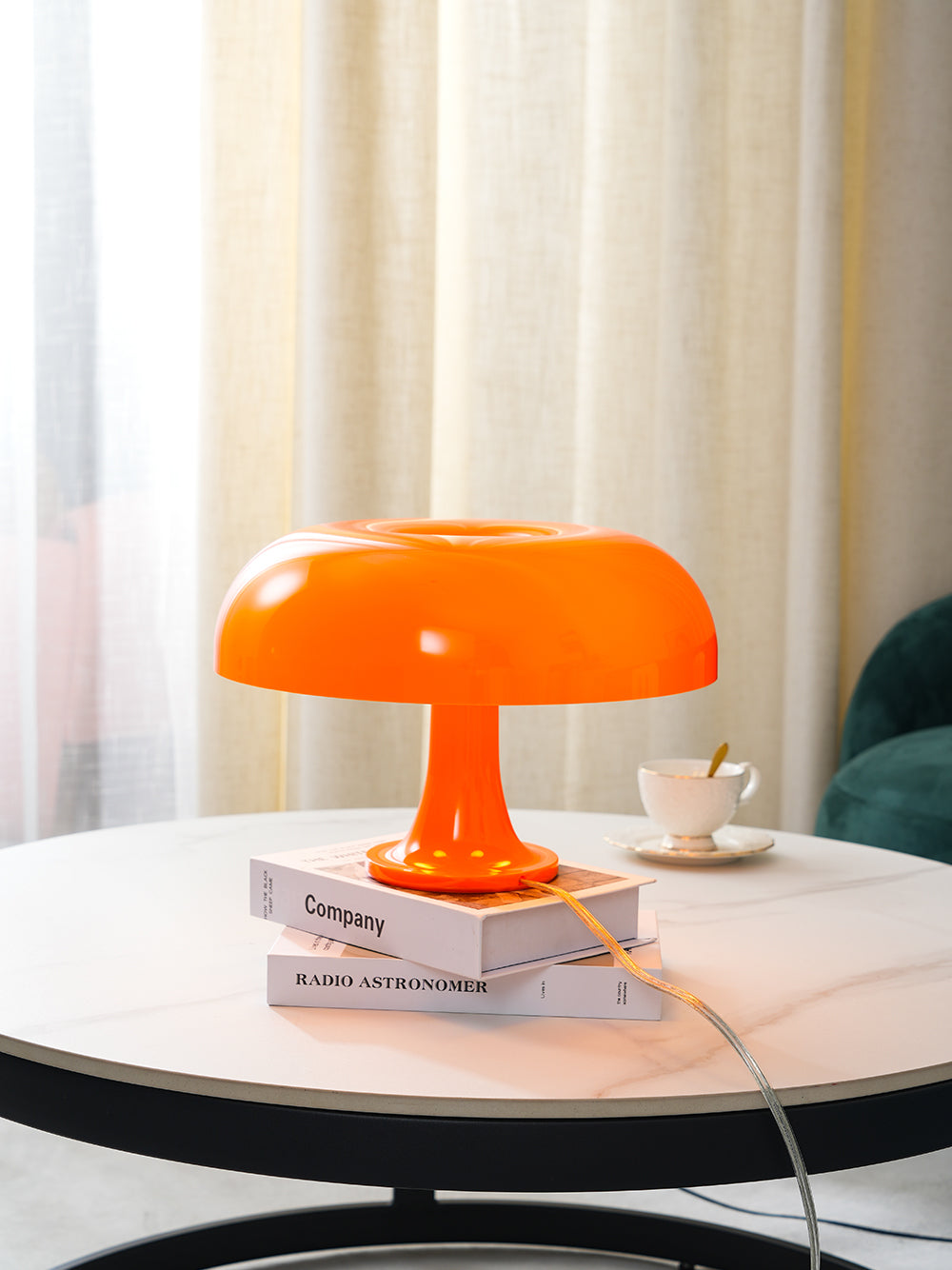 Orange Clérachic table lamp glowing on stacked books and a teacup saucer, with cream curtains behind.