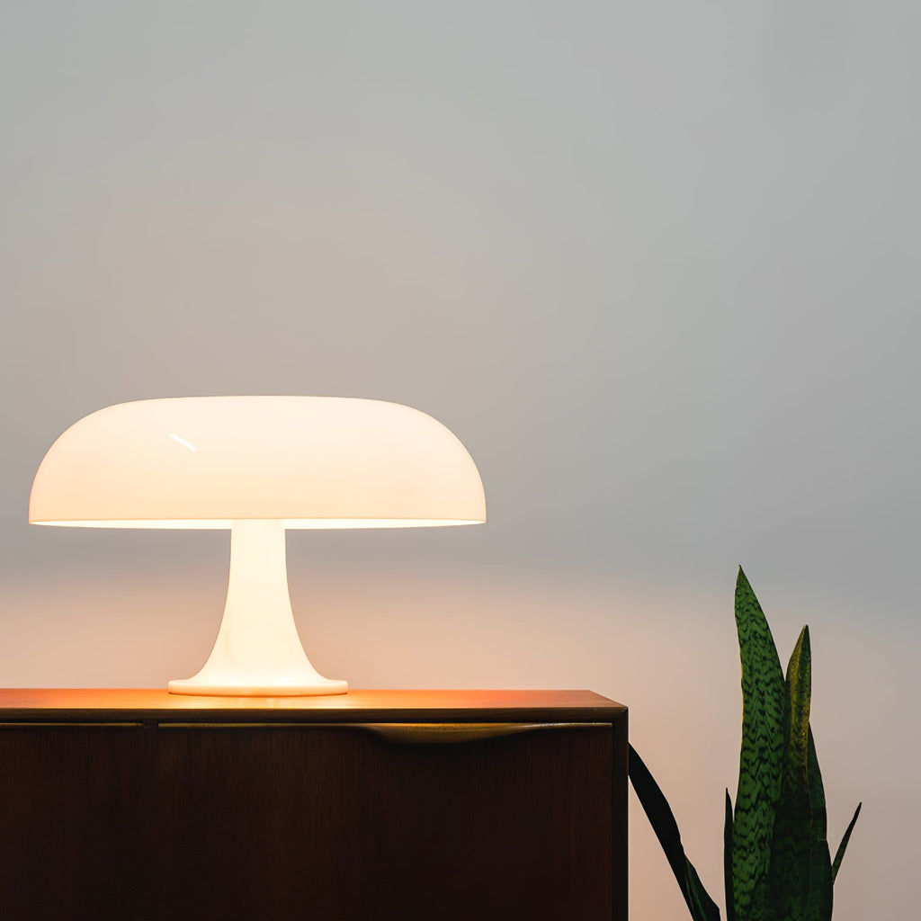 A Clérachic white table lamp giving off warm light on a dark sideboard, with a green plant leaf at the edge.