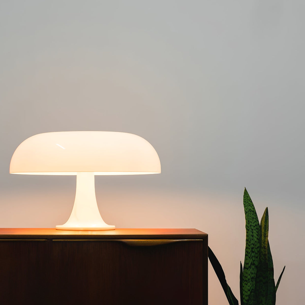 A Clérachic white table lamp giving off warm light on a dark sideboard, with a green plant leaf at the edge.