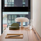 White Clérachic dome lamp on a wooden desk by a tall window, with a notebook, pencil and glass jug beside it.