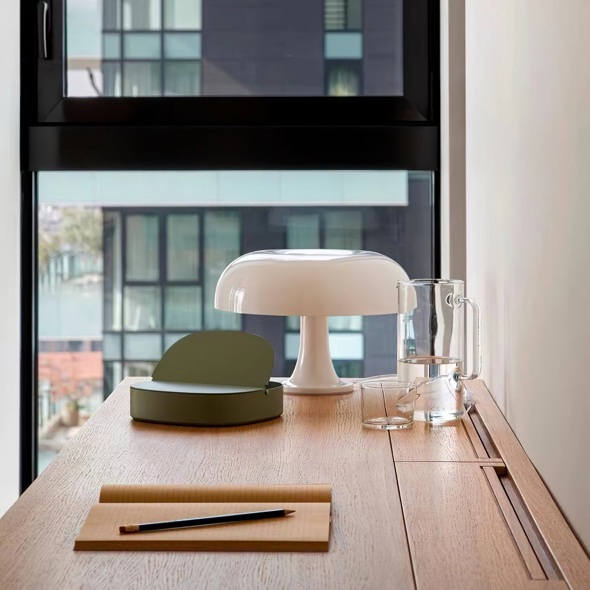 White Clérachic dome lamp on a wooden desk by a tall window, with a notebook, pencil and glass jug beside it.