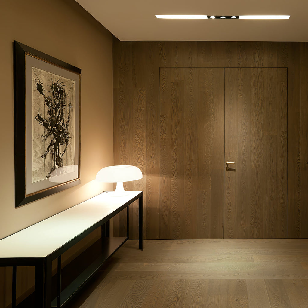 Small white Clérachic lamp glowing on a low bench in a hallway with wood panelling and a large framed print.