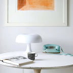 White Clérachic table lamp on a round white table beside a teal telephone, a patterned mug and an open book.