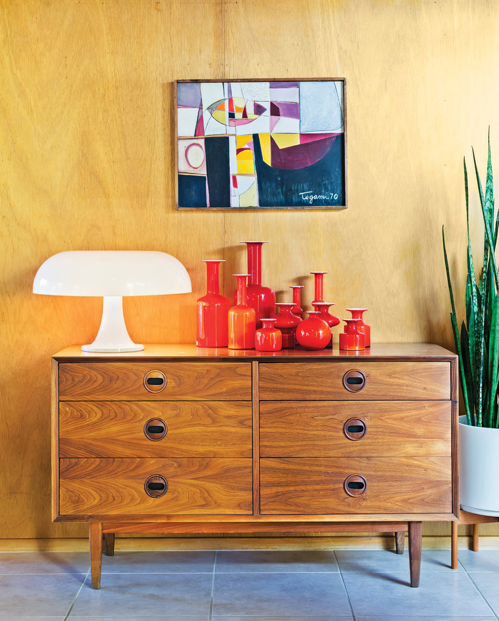 A Clérachic white table lamp on a walnut chest of drawers, with red glass vases and a framed print on a mustard wall.