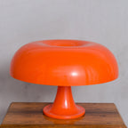 Large orange Clérachic table lamp on a wooden surface against a plain grey wall, its shade seen head on.
