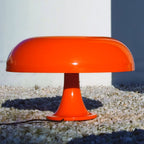 Orange Clérachic table lamp outdoors on pale gravel, with its shade catching the sunlight and a white wall behind.