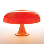 Orange Clérachic table lamp lit so the dome shade glows deep red-orange, on a white background with a warm halo below.