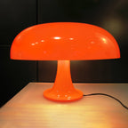 Orange Clérachic table lamp glowing in a dark room, standing on a pale wooden table with a black cord on the right.