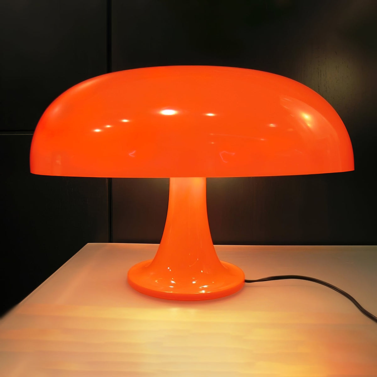 Orange Clérachic table lamp glowing in a dark room, standing on a pale wooden table with a black cord on the right.