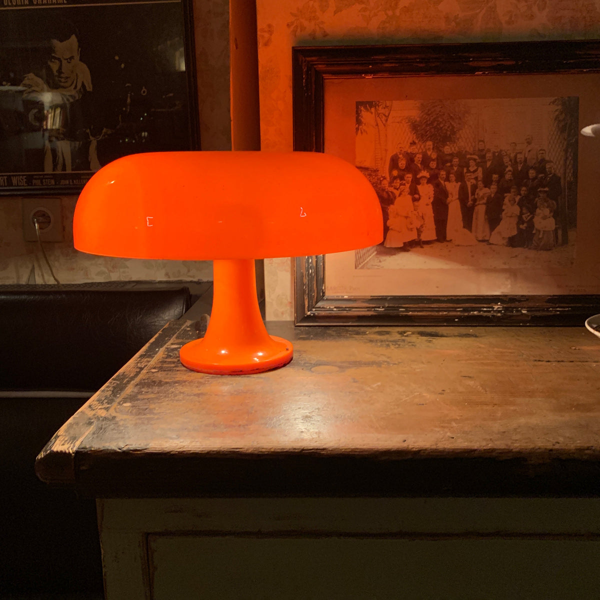 A Clérachic orange table lamp lit on a worn wooden table, with a large framed black-and-white photo behind it.