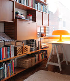 Small orange Clérachic lamp glowing on a white trestle desk in front of a bookshelf, with sunlight through the window.