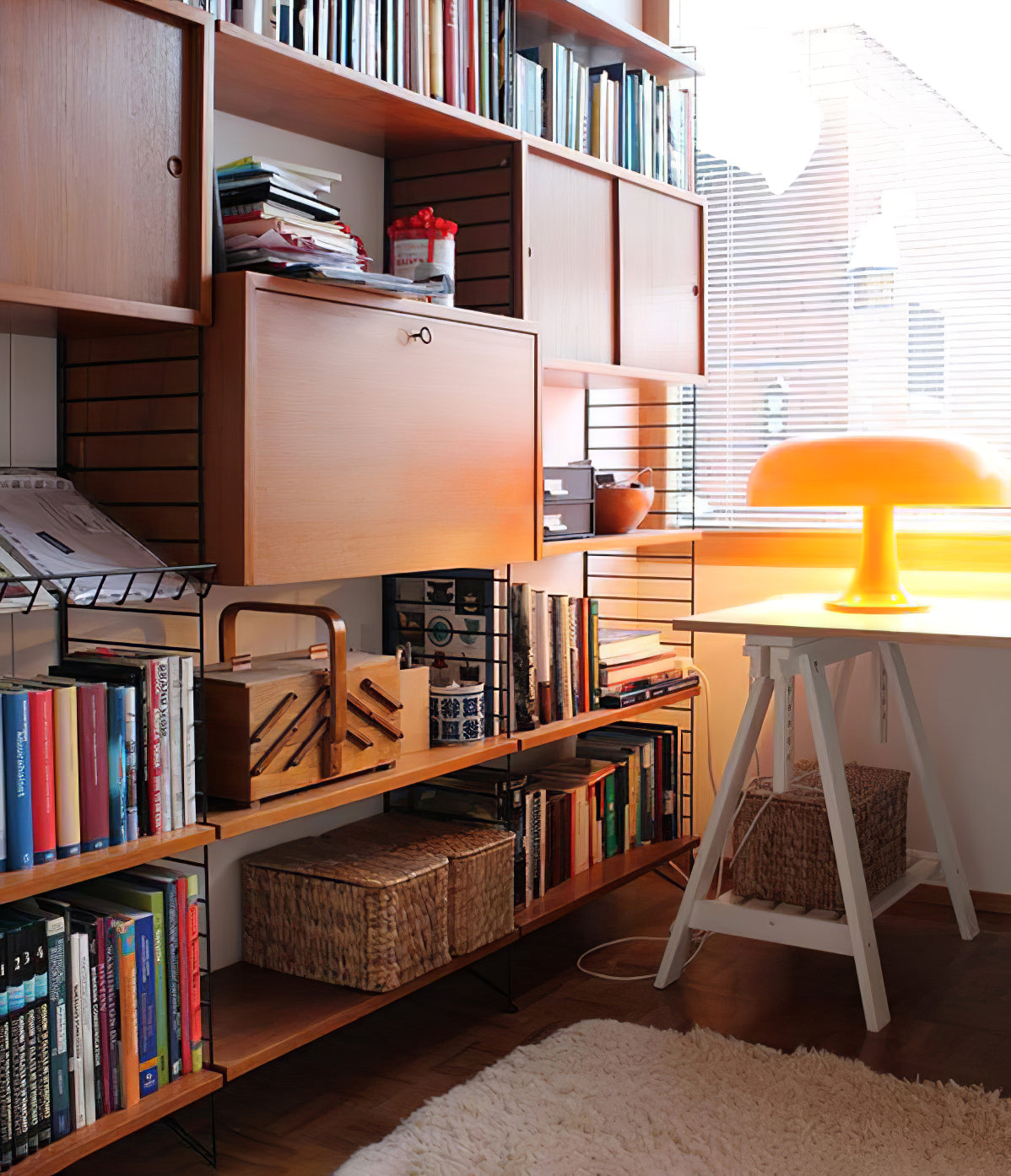 Small orange Clérachic lamp glowing on a white trestle desk in front of a bookshelf, with sunlight through the window.