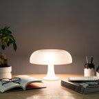 White Clérachic table lamp lit on a wooden desk beside an open book, magazines and a pot of pens, against a grey wall.