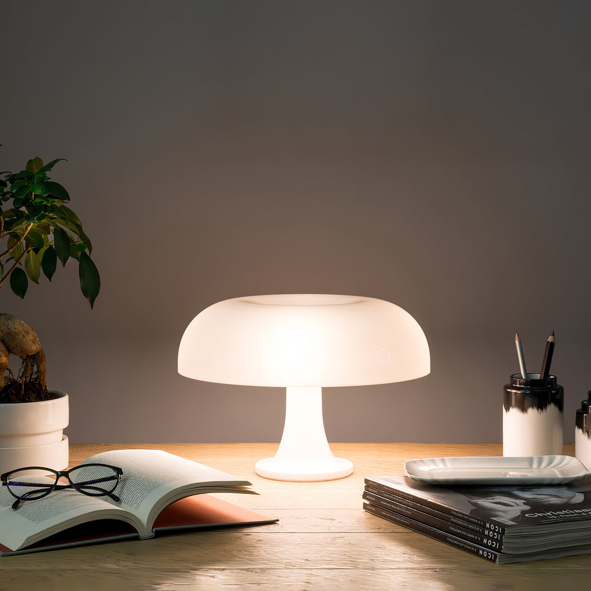 White Clérachic table lamp lit on a wooden desk beside an open book, magazines and a pot of pens, against a grey wall.