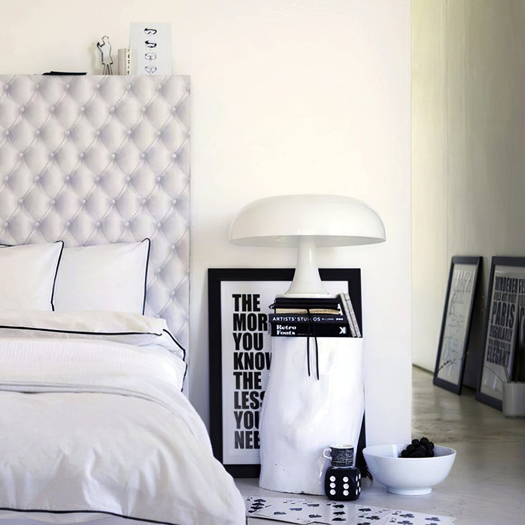 A white Clérachic dome lamp on a stack of books beside a bed with white linen and a black-and-white typographic print.