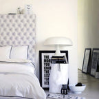 A white Clérachic dome lamp on a stack of books beside a bed with white linen and a black-and-white typographic print.