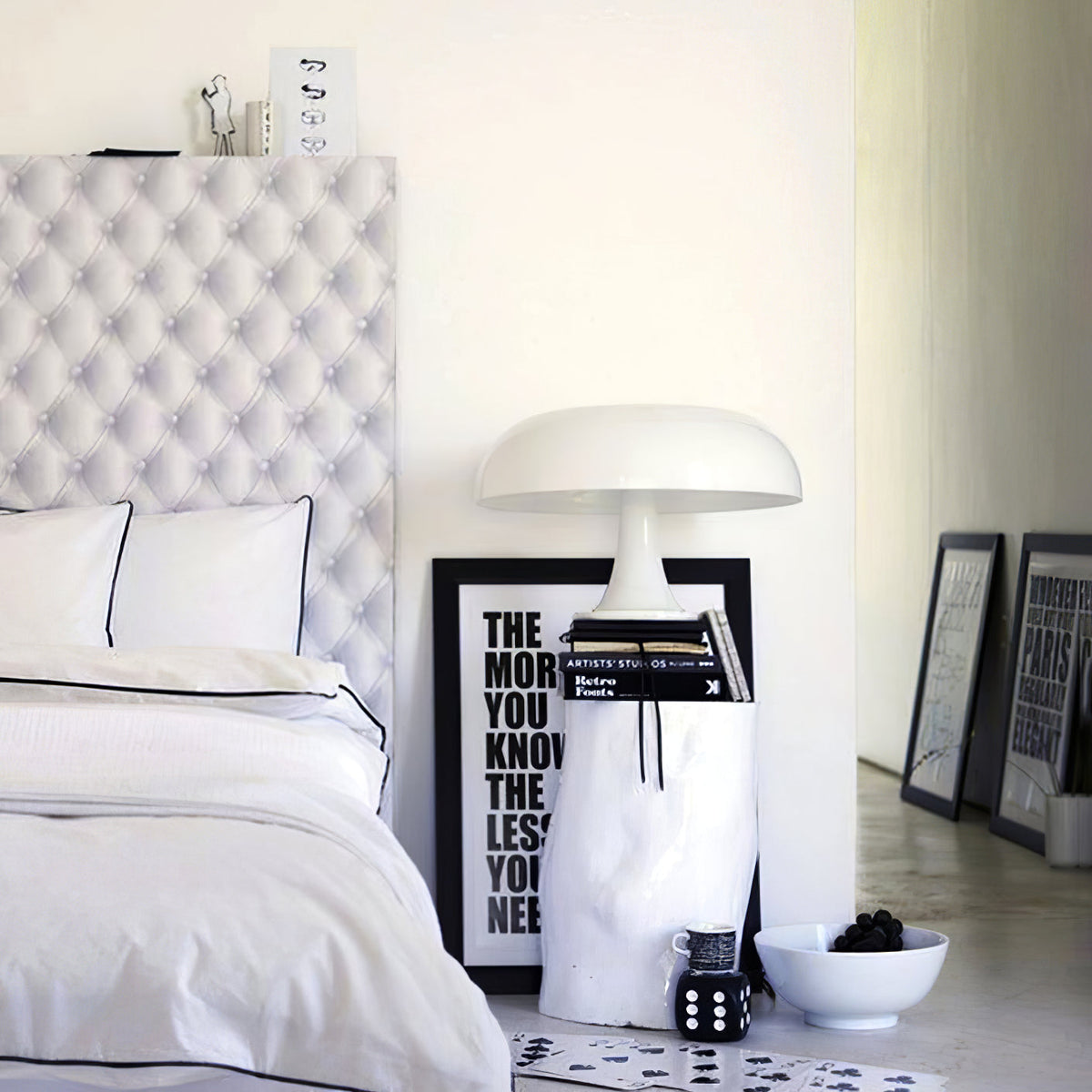 A white Clérachic dome lamp on a stack of books beside a bed with white linen and a black-and-white typographic print.